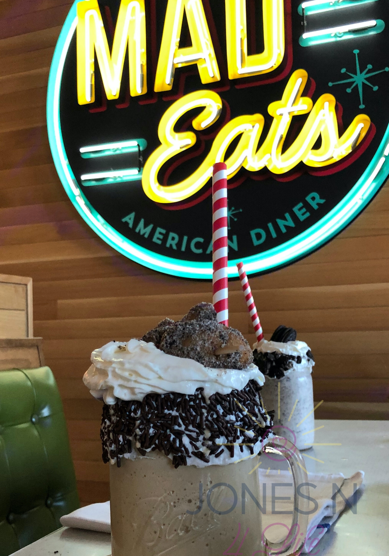 Two chocolate malts sit on a table in restaurant in front of the Mad Eats restaurant sign hanging on the wall in background.